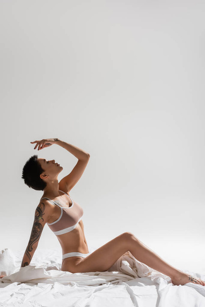 vista lateral da mulher jovem, sexy e encantadora em lingerie bege, com cabelo curto morena e corpo tatuado sentado com a mão acima da cabeça em roupa de cama branca e fundo cinza, fotografia erótica - Foto, Imagem