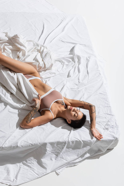 vista superior de mujer impresionante y esbelta en lencería beige, con pelo corto morena y cuerpo tatuado sexy acostado con los ojos cerrados en la ropa de cama blanca en el estudio, fotografía erótica - Foto, Imagen