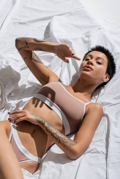 visão de alto ângulo de mulher jovem e apaixonada em lingerie bege, com corpo tatuado sexy e cabelo morena curta deitado em roupa de cama branca e olhando para a câmera em estúdio, fotografia erótica - Foto, Imagem