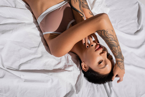 top view of young and charming woman with short brunette hair and sexy tattooed body laying in beige lingerie on white bedding and looking at camera, art of seduction - Φωτογραφία, εικόνα