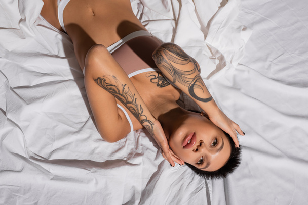 top view of charming and passionate young woman in beige lingerie, with sexy tattooed body and short brunette hair touching face and looking away while laying on white bedding - Foto, immagini