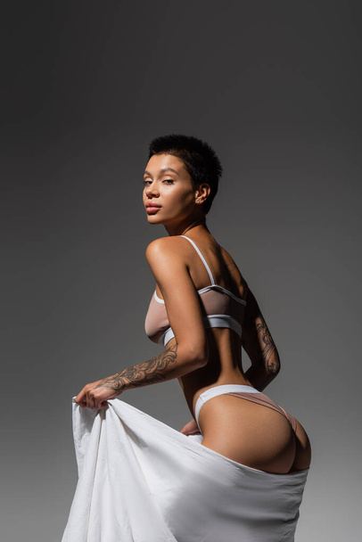 young appealing woman with tattooed body, short brunette hair and sexy buttocks, wearing beige lingerie and looking at camera while posing with white bed sheet on grey background - Foto, afbeelding