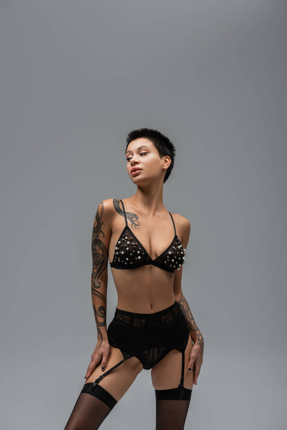 tattooed and expressive woman with short brunette hair and tattooed body posing in bra with pearl beads, lace panties, garter belt and black stockings and looking away on grey background - Photo, Image