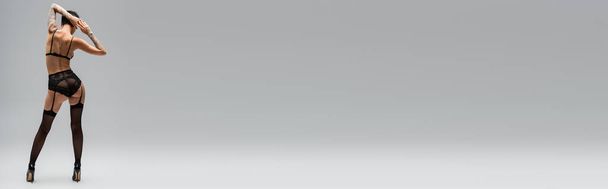 vista posteriore della donna seducente tatuata con corpo sexy e capelli corti bruna in piedi in reggiseno, mutandine di pizzo, cintura giarrettiera, calze nere e tacchi alti su sfondo grigio, banner - Foto, immagini