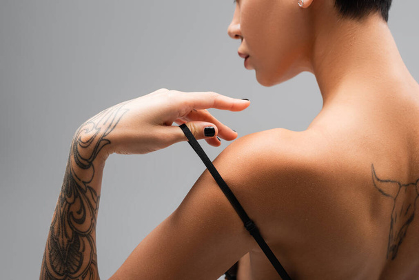 vista parcial de mujer joven y apasionada con sexy cuerpo tatuado tocando correa de sujetador negro mientras posa sobre fondo gris, arte de la seducción, fotografía erótica - Foto, imagen