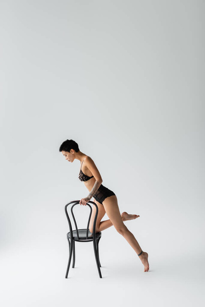 full length of glamorous and tattoo woman with sexy body and short brunette hair ποζάροντας με μαύρη καρέκλα σε σουτιέν με πέρλες και δαντελωτές κιλότες σε γκρι φόντο - Φωτογραφία, εικόνα