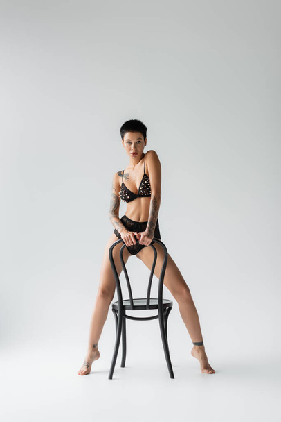 full length of young seductive woman with short brunette hair and sexy tattooed body, in bra with pearl beads and lace panties looking at camera while posing with chair on grey background - Fotoğraf, Görsel