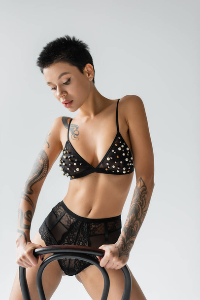 young expressive woman with short brunette hair and sexy tattooed body posing near chair in black bra with pearl beads and lace panties on grey background, erotic photography - Photo, Image