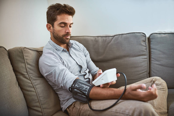 Hypertension, machine and man exam blood pressure in a home sofa or living room for medical or health test. Monitor, reading and person with equipment for chronic disease due to stress. - Foto, afbeelding