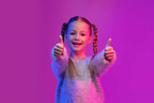 А мне нравится. Счастливая симпатичная девочка-подросток с брейдами в стильном свитере, показывающая большие пальцы вверх и улыбающаяся на цветном фоне при светящемся неоновом свете, малыш рекомендует что-то классное, приятное - Фото, изображение