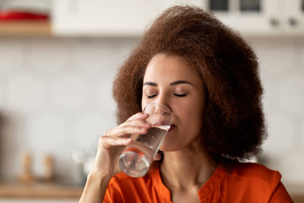 Νεαρή Αφρο-Αμερικανίδα γυναίκα πίνοντας καθαρό μεταλλικό νερό από γυαλί στο σπίτι, διψασμένη μαύρη γυναίκα απολαμβάνοντας υγιεινό ποτό, φροντίζοντας για την ενυδάτωση του σώματος, κοντινό πλάνο shot με ελεύθερο χώρο - Φωτογραφία, εικόνα