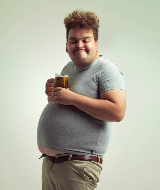 Нет ничего лучше пива после долгого дня. Человек с избыточным весом выглядит довольным, балансируя пиво на животе - Фото, изображение