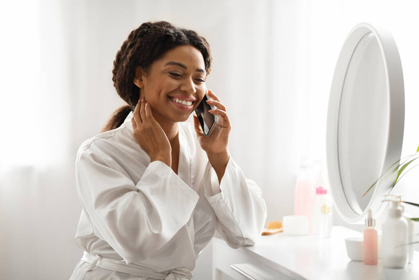 Boldog fekete fiatal nő beszél a mobiltelefon, miközben ül a ruha asztal a hálószobában, gyönyörű mosolygó afro-amerikai hölgy, amelynek kellemes telefonbeszélgetés, miközben a szépség rutin otthon - Fotó, kép