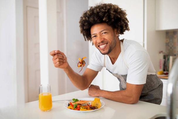 Gourmet casalingo. Uomo afro-americano che si gode il pasto mangiando pesce e verdure cotte sorridendo alla macchina fotografica nella cucina moderna a casa. Guy avendo fatto in casa cena in posa indossando grembiule - Foto, immagini