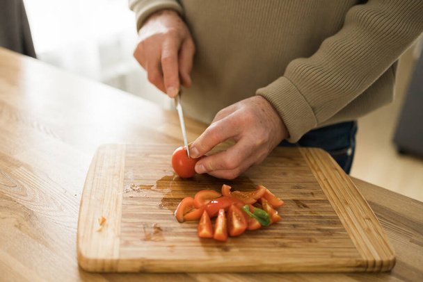 Tuntematon vanhempi mies leikkaa tomaatteja leikkuulaudalla keittiössä - Valokuva, kuva