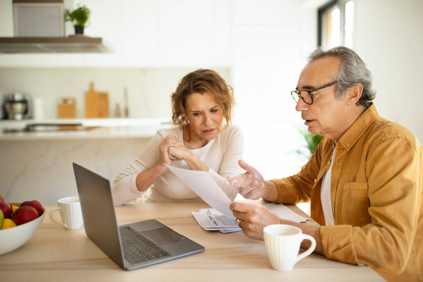 Dotyczące starszych małżonków mężczyzna i kobieta siedzący przy stole we wnętrzu kuchni przed laptopem, sprawdzając rachunki, robiąc liczenia lub pracując razem nad budżetem rodzinnym - Zdjęcie, obraz