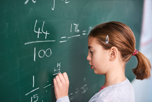I hope this is right. a focussed young elementary school kid writing answers to math questions on a green chalkboard in the classroom - Fotó, kép