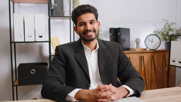 Porträt eines glücklich lächelnden bärtigen indischen Geschäftsmannes am modernen Schreibtisch am Home-Office-Arbeitsplatz, der in die Kamera blickt. Männlicher Hindu im formellen Anzug. Schöner Mann Fernbedienung Online arbeiten auf Laptop - Foto, Bild