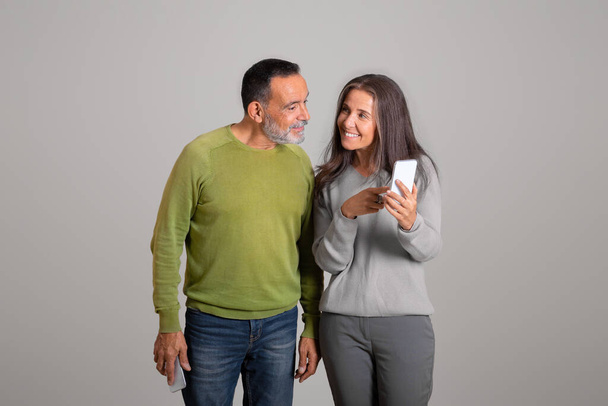 Glad caucasian aged woman shows phone to man, chatting in social network, recommends ad and offer, isolated on gray background, studio. Blog, communication remotely, app banking - Photo, image