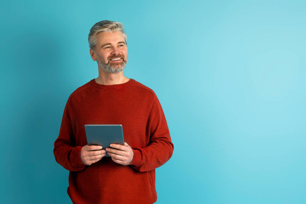 Stock Trading, Financial Planning Online. Happy middle aged man in casual reviewing his investments or managing his finances on mobile app, using digital tablet on blue studio background, copy space - Photo, Image