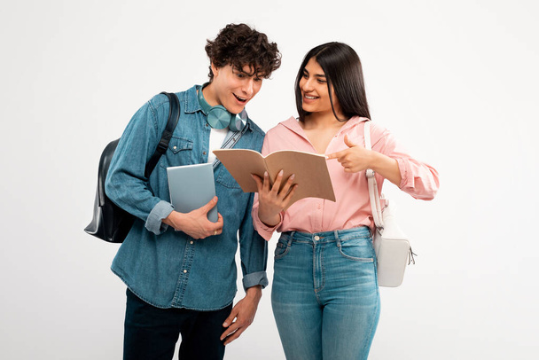College Friends. Cheerful Student Lady Showing Her Workbook To Excited Guy With Backpack, Learning Together Over White Background. Studio Shot Of Enthusiastic Fellow Students Discussing Homework - Фото, зображення