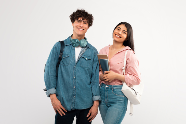 Νεολαία και φιλία. Ευτυχισμένοι μαθητές ζευγάρι ποζάρουν με σακίδιο και εγχειρίδια χαμογελώντας στην κάμερα αγκαλιάζοντας πάνω από το λευκό φόντο του στούντιο. Φωτογραφία του Cheerful College Friends Duo - Φωτογραφία, εικόνα
