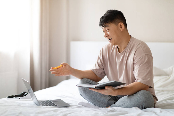 Telecommunication. Positive handsome middle aged chinese man in homewear using laptop in bed at home, holding notepad and pen, have video conference with friends, make plans for weekend, copy space - Photo, Image