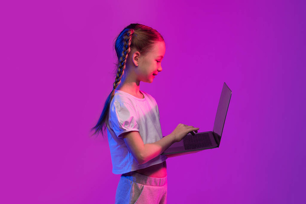 Virtual learning, online education, e-schooling concept. School aged child cute little girl with braids typing on modern pc laptop keyboard over colorful luminous background, copy space, side view - Fotografie, Obrázek