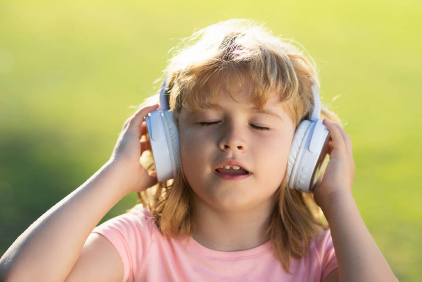 Happy child enjoys listens to music in headphones over green grass background. Funny kid in headphones on summer park. Portrait of kid in headphones listening music with closed eyes - Valokuva, kuva