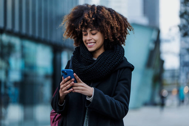 jonge Afro-Amerikaanse zakenvrouw met behulp van mobiele telefoon in de stad van Latijns-Amerika, Latijns-Amerikaanse financiële en caribische mensen met wolkenkrabber achtergrond - Foto, afbeelding