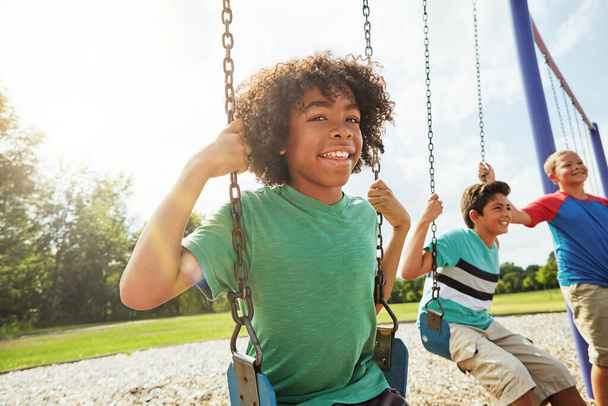 Making it a day of outdoor play. Portrait of a young boy playing on a swing at the park with his friends - Foto, Bild