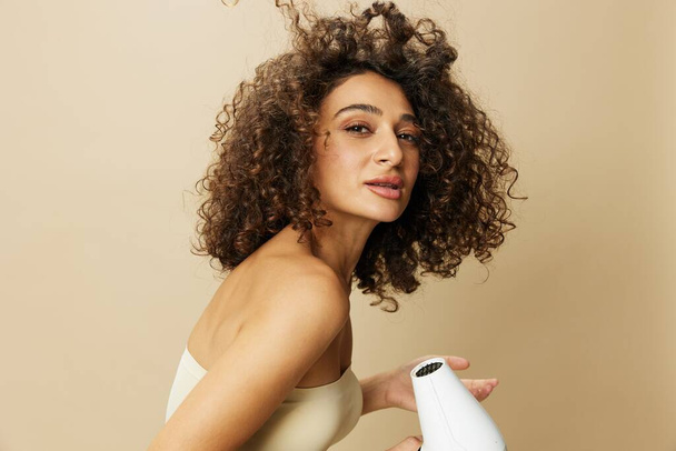 Femme sèche les cheveux bouclés avec sèche-cheveux, maison bat des produits coiffants, sourire sur fond beige. Photo de haute qualité - Photo, image