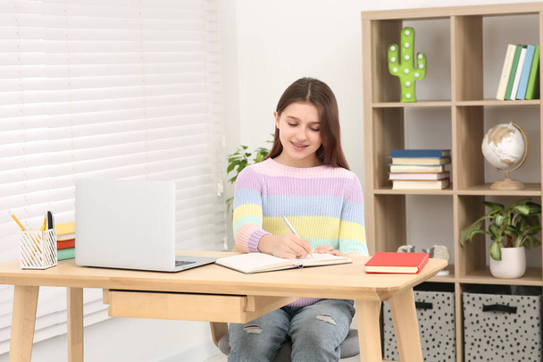 Cute girl writing in notepad near laptop at desk in room. Home workplace - Foto, Imagem