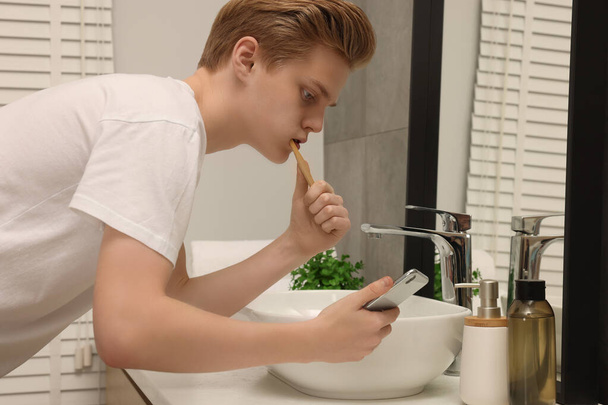 Teenage boy using smartphone while brushing teeth in bathroom. Internet addiction - Photo, Image
