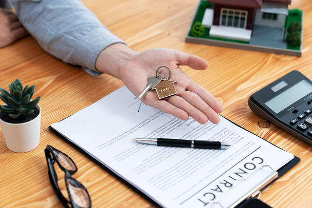 Der Immobilienmakler präsentiert den Hausschlüssel in der Hand über einem Holztisch, umgeben von Taschenrechnern und juristischen Unterlagen, als symbolische Geste für überzeugende Hausverkäufe und Hypothekenangebote. Einheit - Foto, Bild