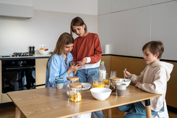 Teen daughter showing new video for social media on smartphone to mom during family breakfast on kitchen. Mother son eating sitting at table. Motherhood, parenting, rising children, mutual support. - Фото, изображение