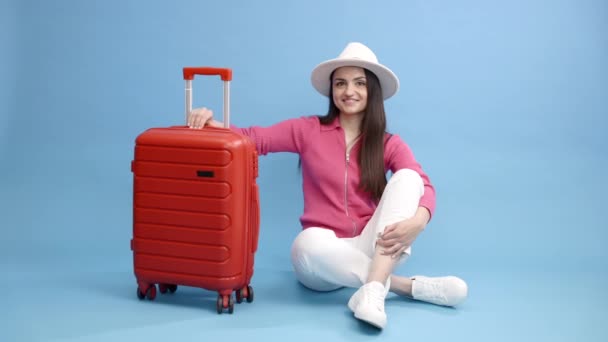 Bavulun başında oturan ve tatillerde mavi arka planda tek başına seyahat eden mutlu genç turist kadın.. - Video, Çekim