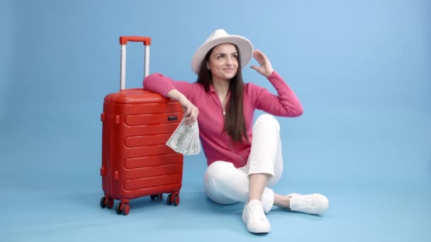 Viaggiatore turista donna in abiti casual e cappello bianco in possesso di denaro e seduto presso la valigia rossa isolato su sfondo blu. Passeggero donna che viaggia all'estero per viaggiare week-end fuga - Filmati, video