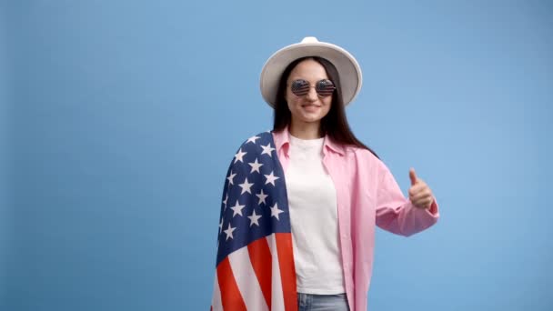 Giovane donna felice sorridente indossa abiti casual e cappello bianco con bandiera americana e mostra un dito su gesto isolato su sfondo di colore blu - Filmati, video