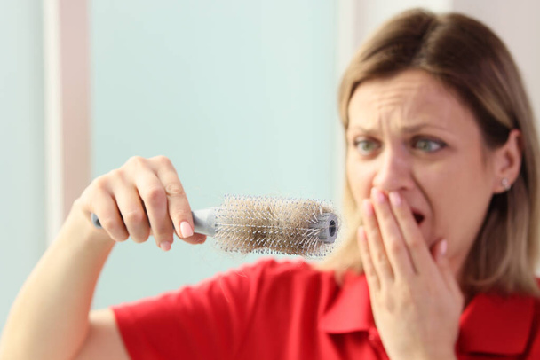 Scared woman holds hairbrush with fallen tangled hair in hand covering mouth. Female person looks upset to discover hair loss at home - Foto, Imagen