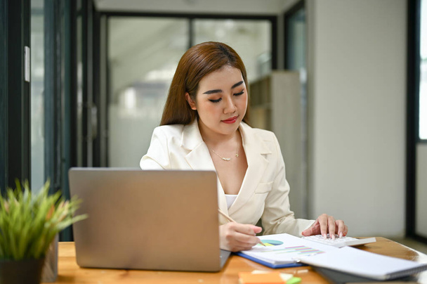 Negocio asiático milenario enfocado y profesional o gerente ejecutivo femenino en un traje blanco que lee y examina informes de negocios en su escritorio en la oficina. - Foto, imagen
