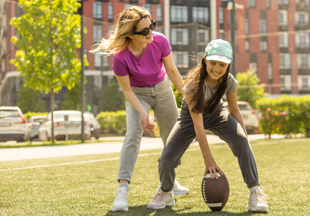 Μητέρα, κόρη, οβάλ καφέ δερμάτινη μπάλα ράγκμπι που παίζει αμερικάνικο ποδόσφαιρο στο πάρκο. Οικογένεια και παιδιά, έννοια της φύσης - Φωτογραφία, εικόνα