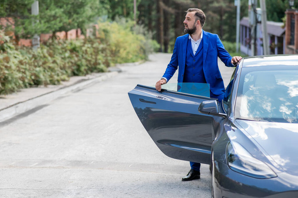 Caucasian bearded man in a blue suit gets out of a black electro car in the countryside in summer - Foto, afbeelding