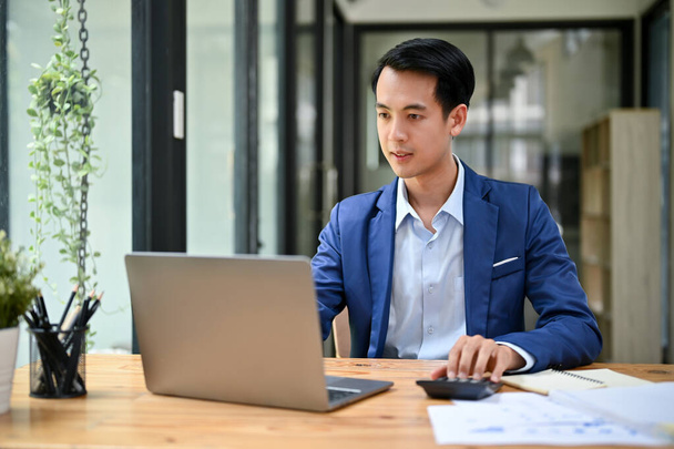 Een knappe en slimme duizendjarige Aziatische zakenman of mannelijke baas in een formeel business suit richt zich op zijn werk op laptop computer in zijn kantoor. - Foto, afbeelding