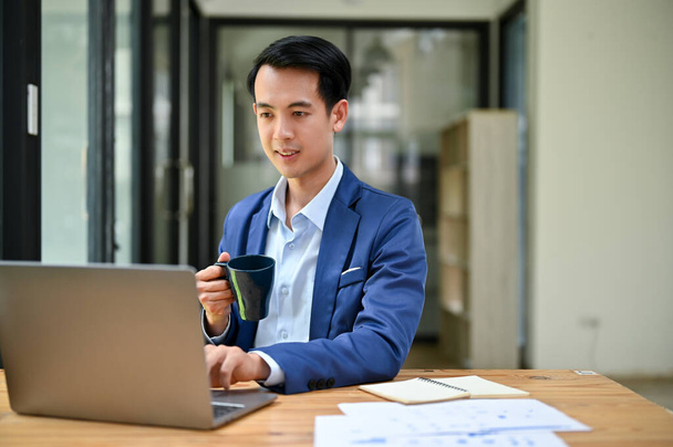 Een knappe en slimme duizendjarige Aziatische zakenman in een formeel zakenpak richt zich op zijn werk op zijn laptop computer terwijl hij koffie drinkt in zijn kantoor. - Foto, afbeelding