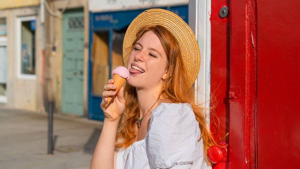 Fiatal, vörös hajú nő szalmakalapban, jégkrémmel a kezében és a szabadban eszi. - Fotó, kép