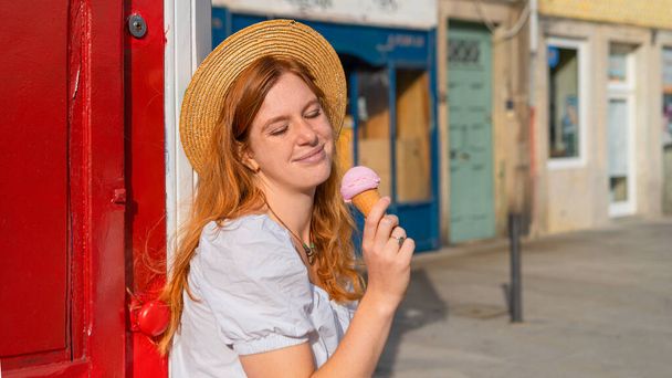 Jovem mulher de cabelos vermelhos adultos em chapéu de palha segurando sorvete e olhando para ele - Foto, Imagem