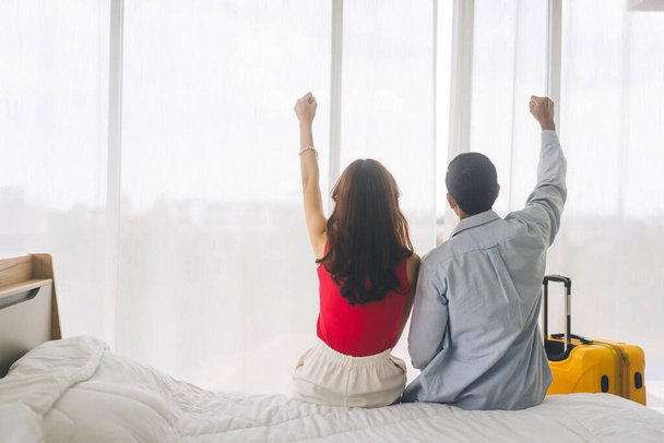 Vista trasera de pareja asiática adulta relajarse brazo estiramiento en el hotel en la cama. La gente viaja de luna de miel por concepto de aniversario. Dormitorio con luz de día de equipaje desde la ventana. - Foto, Imagen