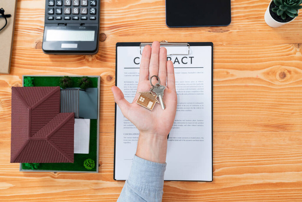 Top view real estate agent presents house key on his hand over wooden table surrounded by calculator and legal documents, as a symbolic gesture for persuasive home sales and mortgage offer. Entity - Foto, Imagen