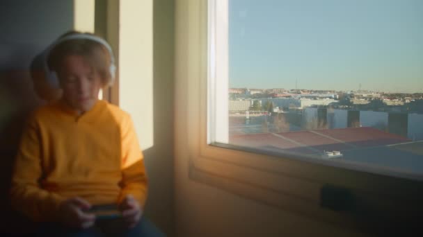 Adolescente de 12 años de edad, se sienta junto a la ventana con vistas a la ciudad, con auriculares blancos, y canta a lo largo de su teléfono durante el amanecer. Enfoque tirando de la ventana al niño al principio de la - Metraje, vídeo
